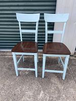 Two wooden bar stools with light blue frames and dark wood seats placed side by side against a corrugated metal door.