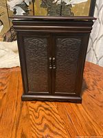 Closed view of wood jewelry cabinet with embossed double doors and metal handles on a wooden table.