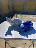 Two ceramic cat figurines placed on a white surface outside. One is white with blue eyes and one small chip, the other is a curled blue cat in sleeping pose.