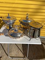 Four stainless steel kitchen items on white table including three chafing dishes and one Bodum fondue set with glass insert and wire stand. All have polished finish with some scratches, lids have various knob handles, some wood handled.