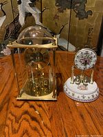 Two mantel clocks on wood grain table, larger brass glass globe Benchmark clock on left, smaller floral battery revolving clock on right.