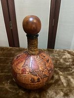 Front angled view of round green glass decanter covered with detailed old-style world map paper, showing North American, Pacific, and parts of Asia with rope detail at the neck and round wooden stopper.