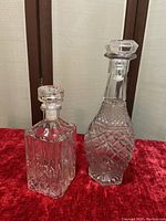 Two clear glass decanters positioned on red fabric with wooden screen background. One square with vertical cuts and faceted stopper, second taller rounded with diamond pattern and ridged stopper.