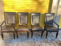 Four dining chairs, two with arms and two armless, brown wood and brown faux leather upholstery with nail head trim, outside on concrete with yellow metal storage doors in background