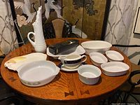View of all items on round wooden table: milk glass pitcher, CorningWare bowls, rabbit platter, gravy bowl and plate, Cambridge spoon, tea cup and saucer