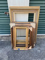 Five wooden frames stacked against a wall, showing various sizes and finishes, some with packing corner protectors