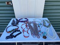Assortment of 11 costume necklaces displayed on a white table, showing various styles and colors including multi-colored seed beads, wooden beads, and pendants.
