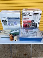 Lot of kitchen items on table includes boxed Black & Decker blender, Instant Pot inner pot box and other items including cat salt and pepper shakers and cookie kit.