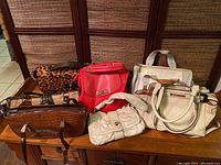 Photo showing six purses on a wooden surface with a background screen. The purses are various colours and materials, including two leopard print purses, a red purse, white purses, and a brown purse.