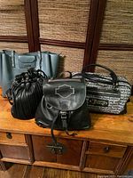Four assorted ladies bags on wooden surface in front of woven backdrop: gray Kenneth Cole tote, Juicy Couture black and gray printed tote, black pleated drawstring bag, and black Harley Davidson leather backpack with flap and buckle.