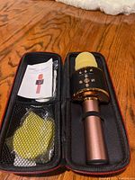 Rose gold handheld karaoke microphone with yellow foam windscreen inside a black zippered carrying case. Mesh pocket contains user manual and yellow USB charging cable.