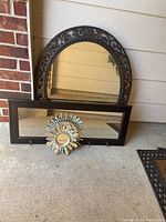 Oval mirror with ornate scrollwork frame behind rectangular mirror with hooks and small sunburst accent mirror in front