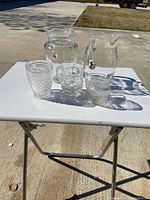 Group photo of two glass pitchers and three glass tumblers on a folding table outdoors, showing the overall lot contents and scale.
