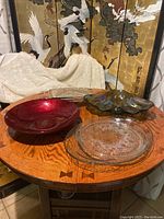 Photo showing assortment of glass and silver serving pieces on round wood table, includes red glass bowl, clear glass platter, iridescent shaped bowl, silver food server.