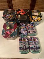 Seven Pokémon collectible tin boxes arranged on a surface showing various Pokémon artwork and windowed lids on some tins.