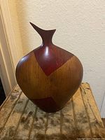 Front view of tall wooden vase with red and brown geometric pattern on a fabric surface, showing scratches and wood grain.