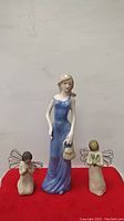 Front view of three porcelain figurines on red surface against white wall, showing girl in blue dress and two small angels