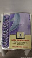 Image showing the packaged Broder fabric shower curtain in purple with matching fabric rings.