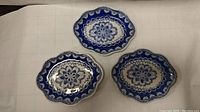 Three hand painted blue and white porcelain plates arranged on a white cloth surface showing floral and geometric designs and scalloped edges.