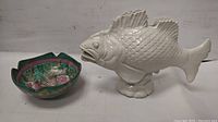 Photo of hand painted porcelain bowl with floral interior and scalloped edge alongside white porcelain fish figurine on base.