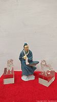 Front view of handcrafted Chinese figurine dressed in blue robes, flanked by two clear crystal tiger statues on cube bases.