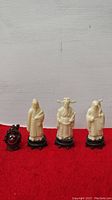 Four Chinese figurines displayed on a red cloth backdrop, three ivory-colored figures on black stands and one smaller dark brown figure in front.