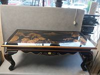 Front view of rectangular black lacquer coffee table with gold detailed landscape artwork on top, curved legs with floral accents.