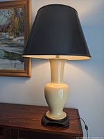Full view of the coffee cream ceramic lamp with black lampshade on a wood surface near a painting.