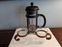 Photo shows the Bodum French press with black frame and lid, and the two clear glass bistro cups with brown handles and rims on a wooden surface.