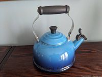Front side image of the blue gradient Le Creuset kettle showing enamel finish and black handle and spout cover.
