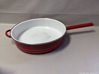 Side angle photo showing the red enamel exterior, loop grip handle, and long handle with white enamel interior surface.