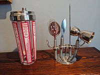 Photo showing the complete set including the glass cocktail shaker with printed red cocktail recipes and the chrome-plated wire holder with bar tools.