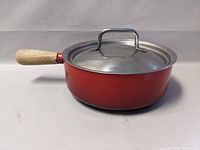 Side view of a red Staub cast enamel pot with wooden handle and steel lid on a white background.