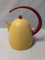 Side profile of a yellow metal tea pot with a curved red handle and round red knob, showing the spout and body shape.