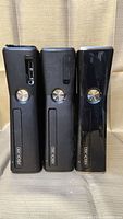 Front view of three Xbox 360 Slim consoles showing Xbox 360 branding and power buttons; one console shows front USB ports.