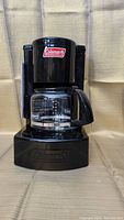 Front view of the black Coleman camping coffee maker showing the glass carafe with measurement markings and black handle. The Coleman logo is visible on the upper part of the device.