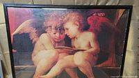 Full view of the framed picture showing two cherubs with wings reading a book, predominantly warm tones. The black frame surrounds the artwork, showing wear.
