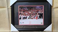 Front view of the framed men's hockey team photo showing the team posing on ice with a crowd in the background. The frame is black with red and black matting.