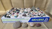 Wide view showing a large collection of over 100 rocks and minerals arranged on a surface, showcasing variety in color and texture from smooth to rough specimens.