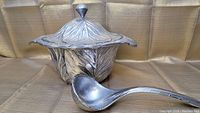 Soup tureen with lid and ladle displayed on fabric backdrop, showing cabbage leaf design and metallic finish.