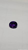 Oval cut deep purple amethyst gemstone on white background showing overall color and facet reflections.