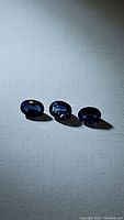 Three loose natural blue sapphires of similar size and shape, oval cut, displayed on a white textured surface with natural light highlighting brilliance and color.