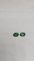 Two natural emerald gemstones placed side by side on a plain white background. The stones have an oval cut and display a vibrant green color with visible inclusions.