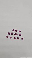 Eleven individually cut rubies laid out on a flat surface. Varied shapes including round, oval, pear, marquise, and emerald. Hot pink to purple color visible in natural lighting.