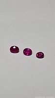 Three oval faceted gemstones displayed on a light background showing the respective hot pink sapphire and red ruby colors, verifying the intense coloration and clarity.