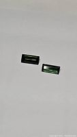 Two rectangular baguette cut green tourmaline stones placed on a white surface, showing deep green color and clear facets.
