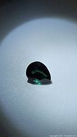 Pear cut blue-green tourmaline gemstone on a light background, showing color and shape under a focused light source.