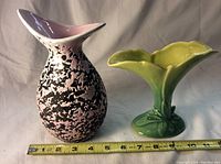 Two vases side by side with measuring tape for scale, showing a mauveish/pink black splatter vase and a light green Beauceware vase shaped with two leaf-like forms.