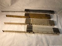 Four ridged folding tape measures laid out flat on fabric background. Show three horizontal and one vertical arrangement with visible measurement markings and signs of wear.