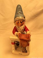 Front view of ceramic gnome figurine named Herbie with tall gray hat, red coat, white gloves, holding a saddle over a white fence with red stripes.
